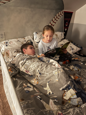 sister | Sanfilippo Syndrome News | a photo of a boy, left, covered up in bed as a young girl reads to him from a book
