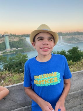get busy living | Sanfilippo Syndrome News | Wearing a bright blue T-shirt and a stylish fedora, Will poses in front of Niagara Falls