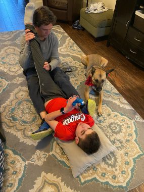 | Sanfliippo News | photo of Will, lying on his back on the floor in a red T-shirt and gray pants. He's on a gray rug with beige and blue designs. His father, in a gray shirt, is stretching one of his legs up toward the ceiling. A dog is lying down beside them. 