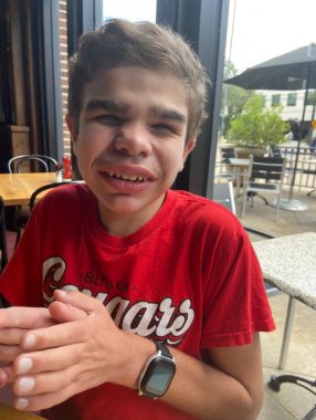 A 12-year-old boy with Sanfilippo syndrome smiles while enjoying lunch at a restaurant. He's wearing a red T-shirt and a smartwatch.