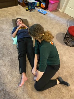 A boy with Sanfilippo syndrome lies on his back on a carpeted floor while a physical therapist kneels over him, working his left foot. The boy has his arms folded on his chest.