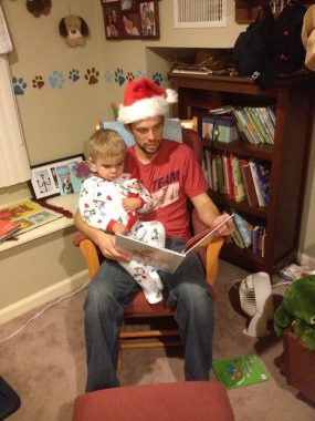 A young father with a Santa hat sits with a 2-year-old boy in pajamas on his lap, with a children's book open. They sit in a children's play room, with a bookshelf full of children's books behind them. The child holds and elf in his arm.