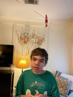 A 12-year-old boy smiles at the camera. Above and behind him in the shot is an upside-down red elf suspended from a red ribbon or string near the ceiling, in what appears to be a living room.