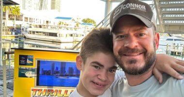 A man and a male teen are seen in head and shoulders, with a yellow information stand and a boats behind them. Both wear light T-shirts; the man wears a gray cap that reads 