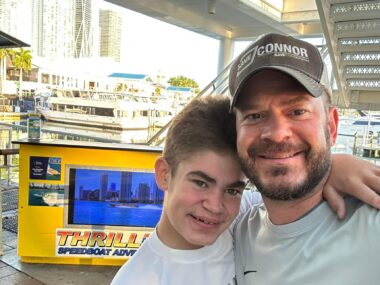A man and a male teen are seen in head and shoulders, with a yellow information stand and a boats behind them. Both wear light T-shirts; the man wears a gray cap that reads 