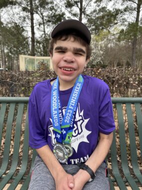 A boy with Sanfilippo syndrome sits on a park bench in a wooded area; he wears a purple T-shirt that is the color to represent Sanfilippo awareness and advocacy, and he has two medals around his neck with the date of 2023. He smiles and looks straight at the camera, with his hands folded on his lap.