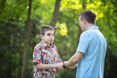 A father and his teenage son stand outside facing each other and holding hands. The father is wearing a light blue short-sleeve shirt while the son is wearing a colorful, patterned button-down, short-sleeve shirt. 