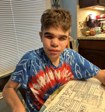 A teenage boy in a blue and red tie-dye T-shirt sits at a kitchen table. 