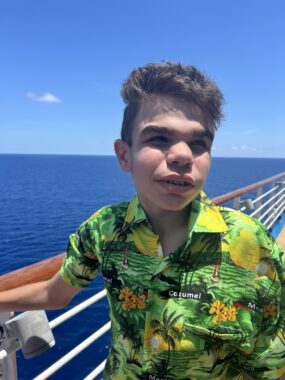 A 13-year-old boy in a stylish yellow and green tropical shirt stands on the deck of a cruise ship. In the background is a vibrant blue sea and cloudless blue sky. A bright sun shines down on the boy's face. 