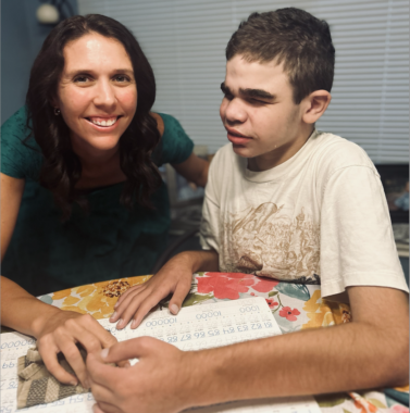 A mom sits at the kitchen table next to her 13-year-old son, Will, who has Sanfilippo. The mom, Valerie, is wearing a green blouse and has freshly curled hair, while Will is wearing a beige T-shirt. Will appears to be working on a school assignment, while Valerie, with one arm around his back and the other resting on the table, smiles at the camera. 