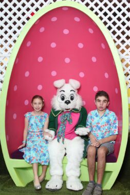 A white Easter bunny, in a dark coat, pink shirt, and colorful tie, sits in a line-green egg with a red background featuring pink dots. White lattice is behind it all. At the bunny's left is a young girl in a bluish-green dress with designs. At the bunny's right is an older boy in shorts and a shirt in the same design as the girl's dress.