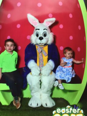 A white Easter Bunny in a dark coat, yellow shirt, and colorful tie sits in the middle of a lime green Easter egg in front of a red background with pink dots. At the bunny's left is a young boy in a green shirt and dark pants; to the bunny's right is a toddler girl in a blue print dress with a pink belt and matching pink flower in her hair.