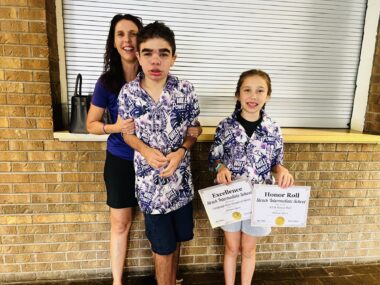 A mom stands with her two children against a brick wall in what could be a classroom. She's just behind her son and holding his arms to support him. Her daughter stands beside them, holding up two award certificates the son earned. The boy and girl are wearing matching purple patterned shirts, while the mom is in a solid purple shirt.