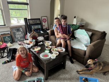 A girl, two boys, and a dog sit near a small couch (or perhaps a love seat) and a coffee table bearing lots of dishes of what appear to be snacks. One boy is embracing the other from behind.