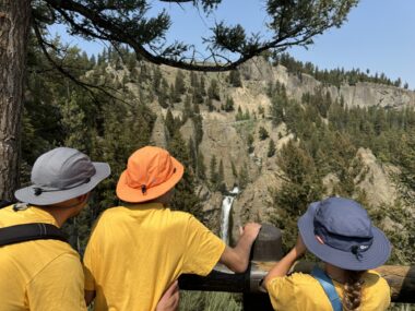 A photo from behind three people shows them looking out at a mountainous ridge lined with pine trees. The three people - an adult and two children - are wearing matching yellow T-shirts and hats to block the sun.