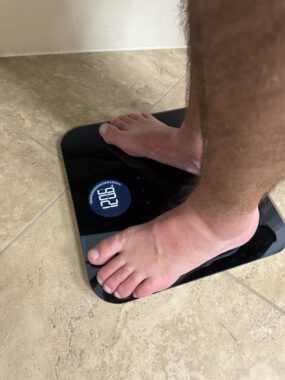 A close-up of a man's feet standing on a digital scale. The scale reads 120.6 pounds. 