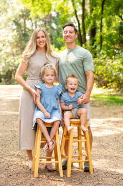 A professional-looking photo shows a family of four posing together on a wide dirt road in the woods. The parents are standing side by side, with the mom in a long khaki dress and the dad in a short-sleeve green shirt. In front of them, sitting on identical wooden stools, are a young girl, maybe 4 or 5 years old, and a boy, maybe 1 or 2 years old. The girl, who has Sanfilippo syndrome type B, is wearing a blue dress, and her brother has on a blue shirt and khaki shorts. The trees behind them are tall and glowing in the sun, and all four family members are smiling at the camera.