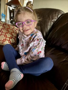 A young girl, about 5 years old, sits cross-legged on a couch at home. Her blond hair is styled in double buns, and she's wearing pink-framed glasses, a pink shirt featuring "Toy Story" characters, blue leggings, and striped socks. She's smiling happily at the camera.