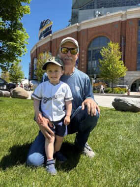 A man with a white cap, glasses, a blue shirt and jeans crouches in the grass, his arm around a toddler standing next to him. The boy wears blue shorts and a white Brewers shirt.