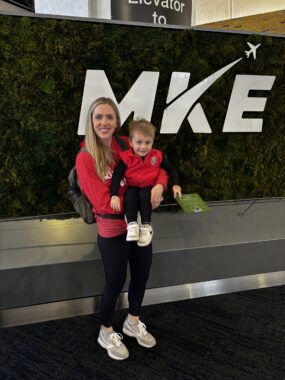 A woman with long blond hair, a backpack, a red top, and black pants holds a blond-haired toddler in a red vest. She's standing before a large black sign reading "MKE," with the top of the K extending into an airplane. Above it you can see part of another sign reading "Elevator to."
