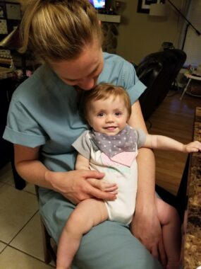 A mother dressed in hospital scrubs holds her baby girl on her lap and looks down on her. The baby smiles and looks at the camera.