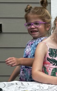A young girl with blond hair, its pair of pigtails tied into buns, is in front of a gray house. She wears pink glasses and what seems to be a floral-print, sleeveless outfit. Beside her appears to be another girl.