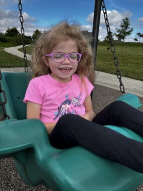 A young girl with lots of blond hair, pink glasses, a pink T-shirt and black pants smiles as she sits in a large, green swing. The outdoors setting looks fairly bare and expansive behind her, though there's a gray path cutting through it.