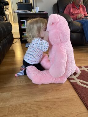 A 2-year-old girl in a polka dot shirt kisses a giant pink pig stuffed animal while both are sitting on the hardwood floor of a home. Someone can be seen sitting on the couch in the background.