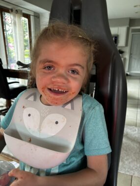 A 5-year-old girl smiles happily after a meal. She's wearing a silicon bib around her neck, and remnants of her lunch are visible around her mouth.