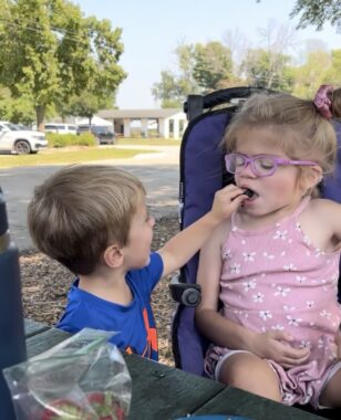 A 5-year-old girl wearing a pink dress and glasses sits in a purple chair outdoors. Her younger brother stands next to her and holds a blueberry up to her mouth for her to eat 