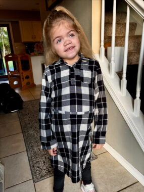 A young girl, maybe about 5 years old, stands in a hallway in her home wearing a black-and-white plaid dress. Her arms are by her sides and her head is tilted at someone to the right of the camera.