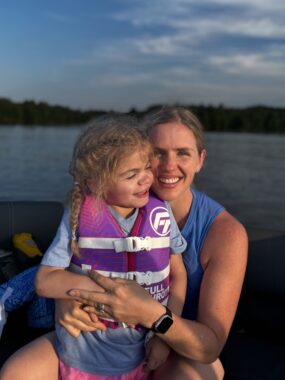 A mom and her daughter smile for a photo near a lake. The girl, maybe about 5 years old, is wearing a life jacket and has her hair in braids. They are illuminated by sunlight, but the background is darker, as if they're catching the last rays of the sun before it sets.