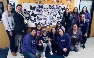 A group of adults is shown gathered around a child. Most are wearing purple, and there's a banner behind them covered with purple butterflies. They appear to be gathered in a school hallway.