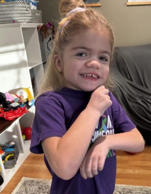 A girl wearing a purple World Sanfilippo Awareness Day T-shirt is seen smiling.