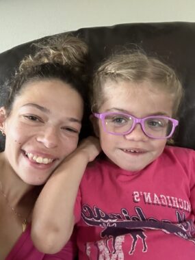 A woman in her 20s takes a selfie with a young girl, maybe 5 or 6 years old. Both are wearing pink and appear to be sitting side by side on a couch.