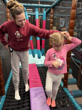 A woman walks beside a young girl in an indoor playground. She's holding one of the girl's hands to help her balance on the black netting they're walking across.