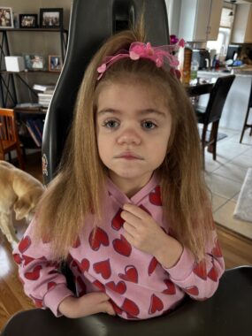 A young girl is dressed up for Valentine's Day by wearing a pink sweatshirt with red hearts and a pink flower headband. She appears to be in an office or living space of a large home. 
