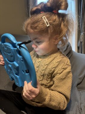 A young girl sits on a couch at home holding a blue tablet. We can't see what's playing onscreen, but she is engrossed in it.