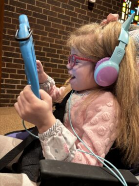 A young girl wearing glasses and headphones laughs while holding up a blue tablet to watch a show. She appears to be sitting at the end of a row of seats in a church, with a brick wall and part of a stained glass window behind her.