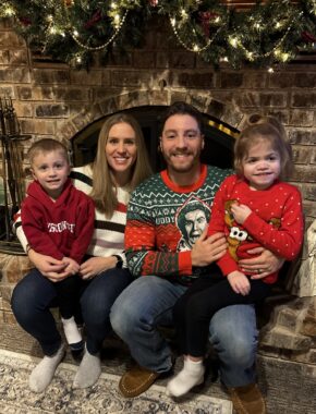 A family of four sit in front of a fireplace hung with Christmas decorations.
