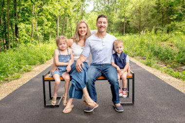 A family of four — mom, dad, and two young children — sit side by side on a bench for a family portrait. The bench has been placed in the middle of a pathway, and they're surrounded by trees and foliage.