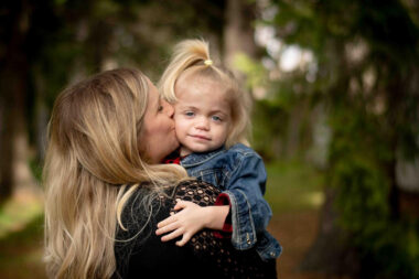 A mother holds her 2-year-old daughter in her arms and kisses the girl's cheek. The girl is hugging her mom and smiling softly at the camera.