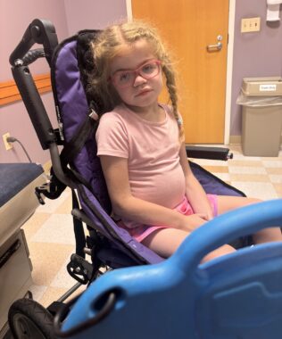 A young girl with her hair braided sits on a walker in a doctor's office.