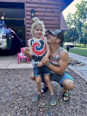 A man kneels beside a young girl outside in the driveway of a home. Both are wearing red, white, and blue clothing, and the girl is holding a sparkler in her right hand.