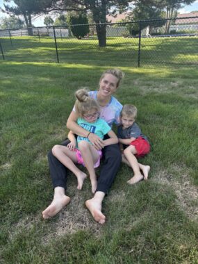 A mom sits outside on a grassy lawn with her two children. Her daughter sits in her lap while her son curls up on her left.