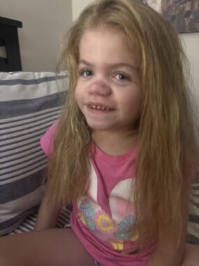 A 6-year-old girl with Sanfilippo syndrome sits on a bed and smiles at the camera. Her hair is long and straight, and she's wearing a pink shirt.