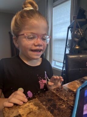 A 6-year-old girl laughs at a tablet propped up on the counter in front of her. She's wearing a black T-shirt, glasses, and has her hair in a bun.